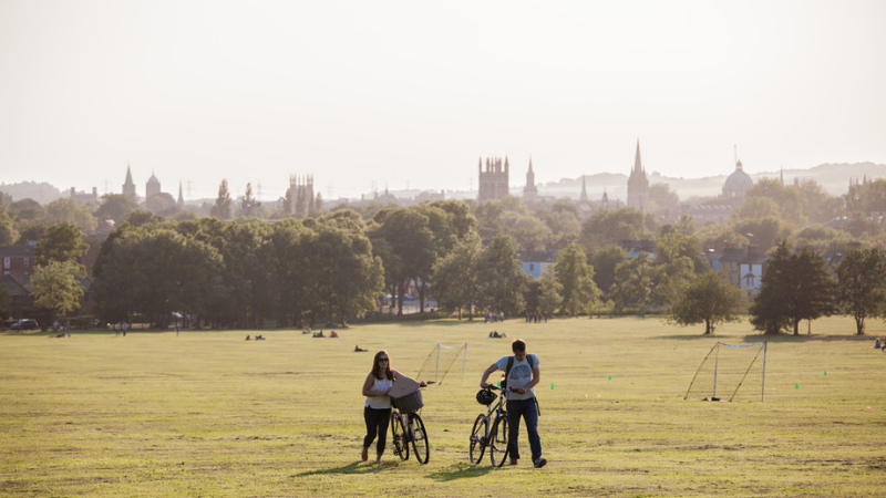 Two students walk beside their bikes through South Park, the Oxford skyline is behind them